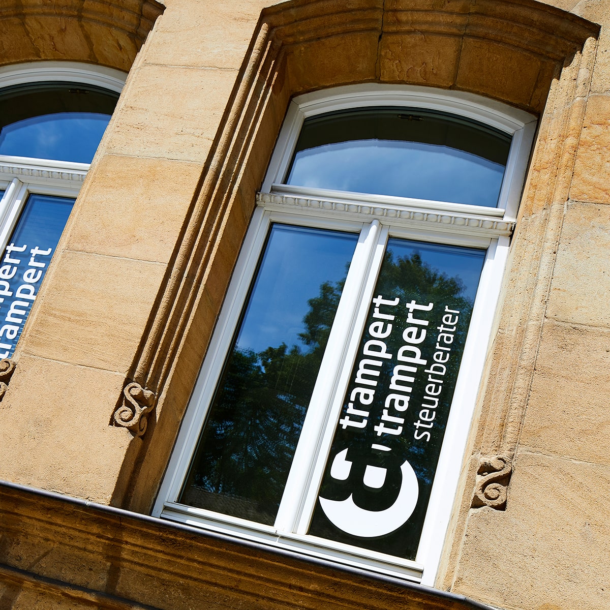Teaser Quadrat verlinkt, trampert & trampert steuerberater Fensterbeklebung Logo weiß, Altbau Sandsteinmauer, Aufnahme dynamisch schräg 