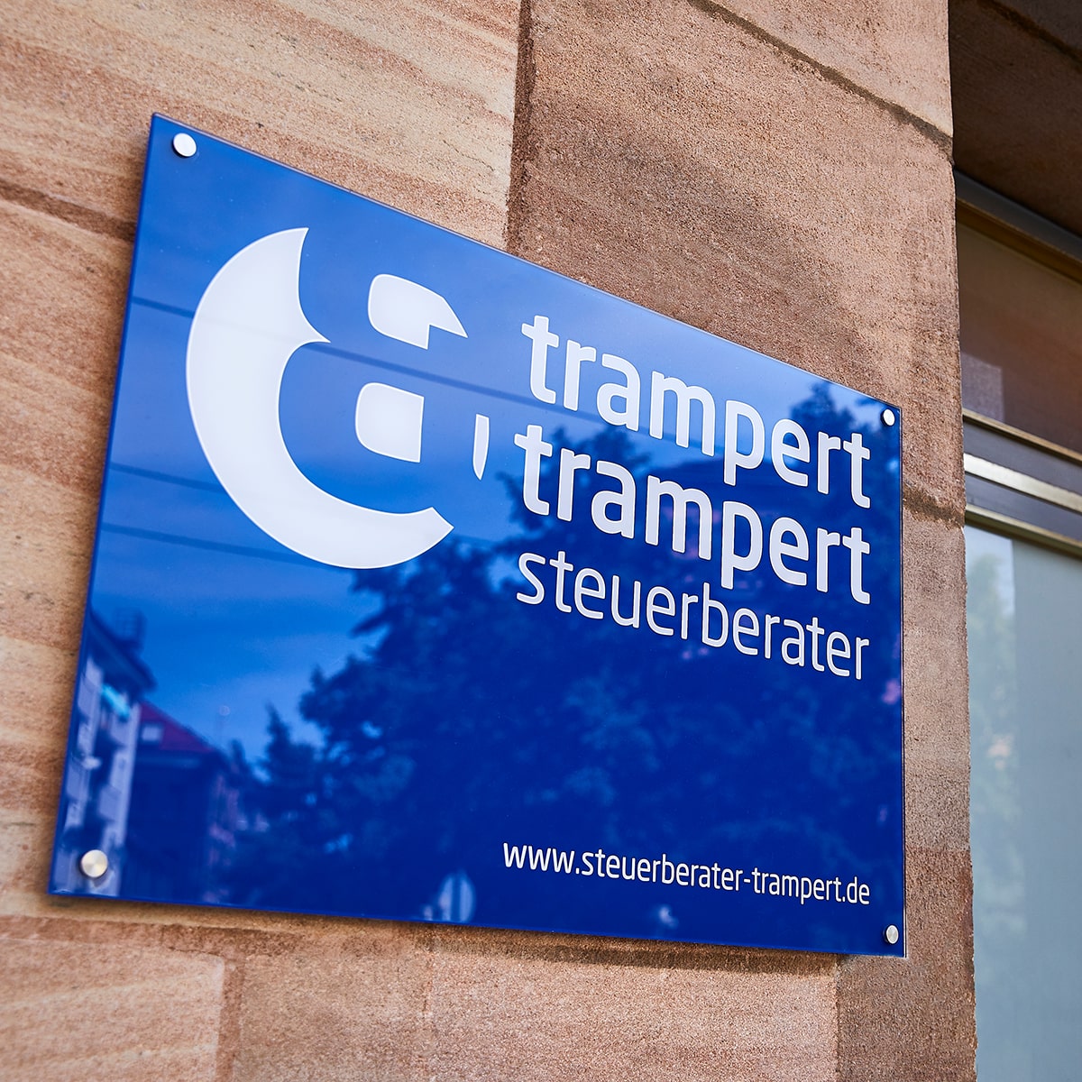 Teaser Quadrat verlinkt, trampert & trampert steuerberater dunkelblaues Firmenschild Plexiglas mit Logo und Webadresse in Weiß, an Hauseingang auf Sandsteinwand montiert, schräg aufgenommen