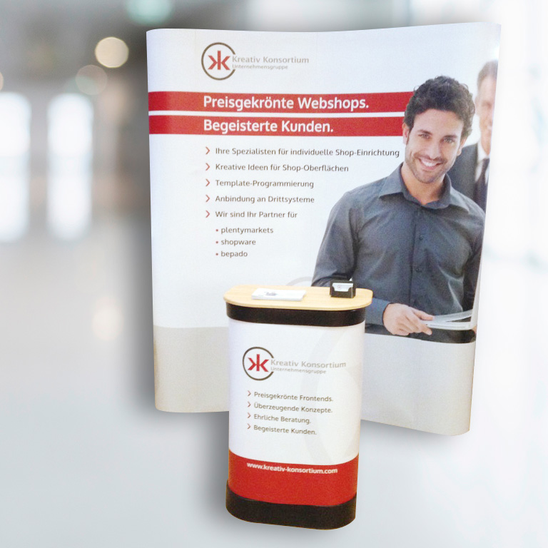 Teaser Quadrat Messeausstattung; mobiler Messestand Kreativ Konsortium GmbH, halbrund mit Logo, Motiv und Text, im Vordergrund ovaler Beratungsdesk; gestaltet mit Logo und Text; Farben Weiß, Hellgrau, Rot